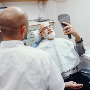 imagem de um paciente idoso vendo no espelho o resultado da reabilitação oral ao lado do seu dentista