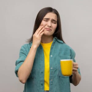 Mulher de pé, vestindo camisão verde e camiseta amarela por baixo, com mão direita apertando o dente, demostrando dor, e na mão esquerda caneca amarela.